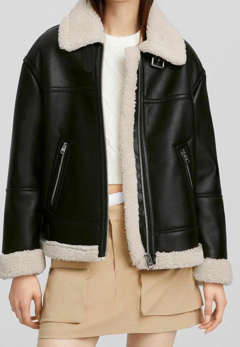 Kvinna som bär en svart konstläderjacka med krämfärgad teddykrage, manschetter och kant, ihop med en vit stickad tröja och en beige kjol med fickor.