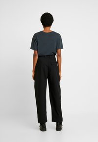 Dunkelgraues kurzärmliges Shirt, kombiniert mit hochtaillierten schwarzen weit geschnittenen Hosen und schwarzen, hohen Stiefeln, von hinten betrachtet.