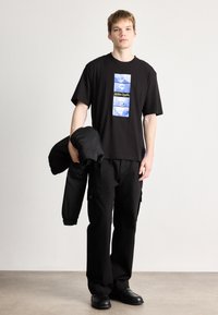 Μαύρο γραφικό T-shirt με μπλε σχέδια και κείμενο, συνδυασμένο με μαύρο cargo παντελόνι και μια μαύρη μπουφάν, κρατημένο στο ένα χέρι.