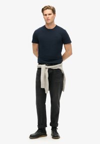 Mand står med hænderne bag ryggen iført en marineblå t-shirt, sorte jeans, sorte støvler og en lys grå sweater bundet om livet foran en hvid baggrund.