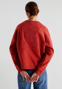 Rode sweatshirt met ronde hals, met een textuurdesign van een glinsterende doodskop en gekruiste beenderen op de achterkant. Het materiaal is een zachte katoenmengsel met geribbelde manchetten.