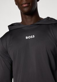 Huppari, jossa on huppu, mustasta sileästä kankaasta ja valkoinen "BOSS"-logo rinnassa. Ominaisuuksiin kuuluu yksinkertainen ja virtaviivainen muotoilu.