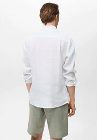 Camisa de lino blanca con mangas largas dobladas, combinada con pantalones cortos verde claro. La camisa presenta un collar clásico y un yugo en la parte posterior.