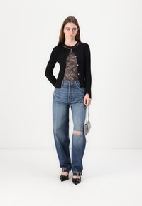Cardigan nero a maniche lunghe sopra una blusa in pizzo trasparente, abbinato a jeans blu a vita alta con ginocchio strappato e scarpe nere con punta.