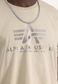 Camiseta de algodón beige con un logo "A" gris brillante y el texto "ALPHA INDUSTRIES, KNOXVILLE, TENNESSEE, U.S.A." Collar de cadena plateada redonda.