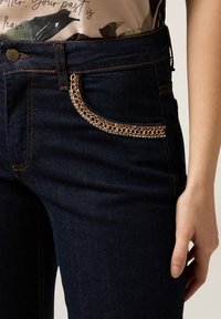 Dark blue denim jeans with orange stitching and a gold chain detail along the front pocket, worn with a beige top with floral print.