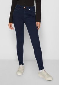 Jeans alta vita di colore blu navy con design aderente, bottone di rame e tasche anteriori. Indossati con sneakers di colore chiaro.