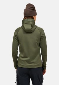 Asmuo, vilkintis alyvuogių žalios spalvos džinsinį švarką su „Peak Performance“ tekstu ant gobtuvo ir rudą vilnonę kepurę, matomas iš už nugaros.