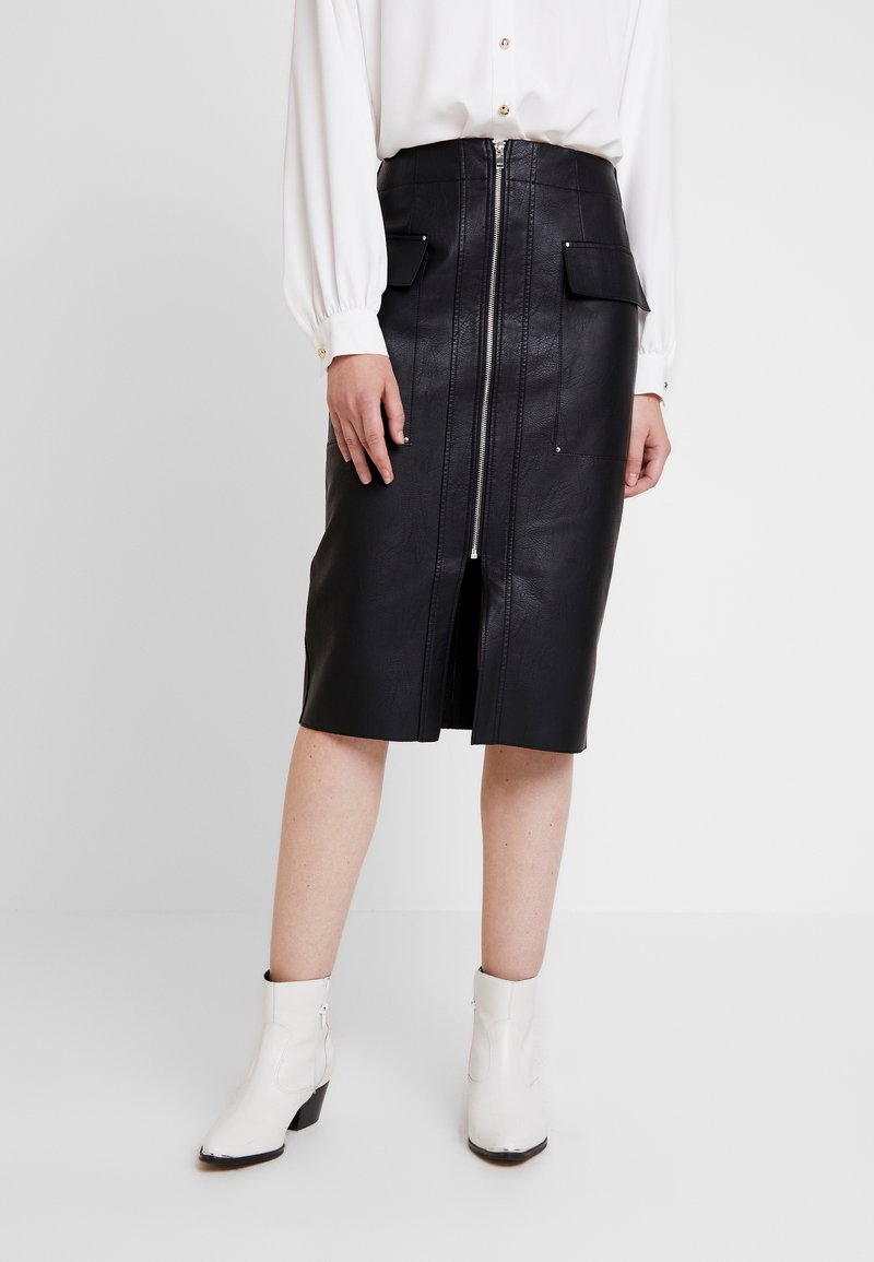 Jupe crayon en similicuir noir avec une fermeture éclair frontale, deux grandes poches plaquées, et un ourlet longueur genou. Portée avec des bottines blanches.