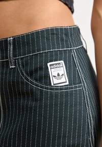 Pantalons en denim vert foncé avec des rayures blanches, présentant une étiquette blanche Adidas bien visible sur la poche arrière et des détails de couture contrastants.