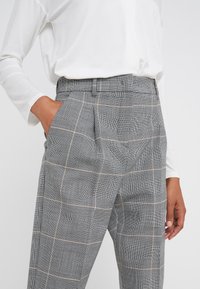 Grijze geruite broeken hebben een hoge taille, zijzakken en een relaxte pasvorm, gemaakt van een gestructureerde stof met fijne zwart-witte patronen.