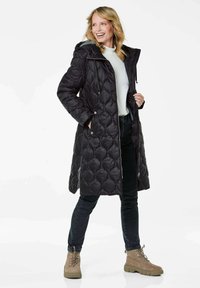 Schwarzer gesteppter langer Mantel mit Kapuze, ausgestattet mit einem Reißverschluss, seitlichen Taschen und dekorativen Stichen; kombiniert mit dunklen Jeans und braunen Stiefeln.