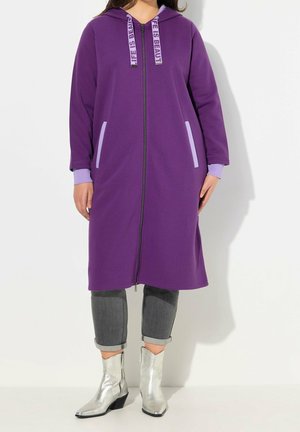 Vrouw draagt een lange paarse zip-up hoodie met "Life is Beautiful" veters, grijze opgerolde spijkerbroek en zilveren enkellaarsjes.