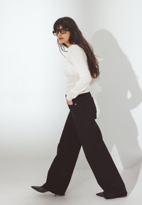 Vit långärmad topp med genomskinlig textur, tillsammans med högmidjade svarta vida byxor och spetsiga svarta klackar. Mörka solglasögon fulländar looken.