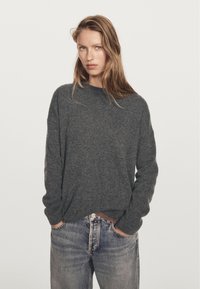 Frau mit langen blonden Haaren, die einen dunkelgrauen Strickpullover und hellblaue Jeans trägt, steht mit den Händen in den Taschen vor einem schlichten Hintergrund.