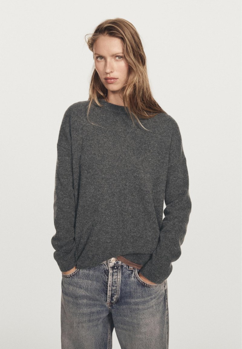 Frau mit langen blonden Haaren, die einen dunkelgrauen Strickpullover und hellblaue Jeans trägt, steht mit den Händen in den Taschen vor einem schlichten Hintergrund.