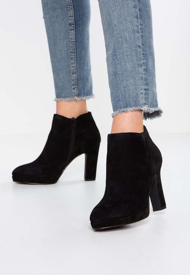 Bottines en daim noir à talons blocs portées avec un jean bleu court à ourlet effiloché, debout sur un sol blanc.