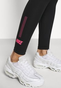 Svarta leggings med en rosa rutdesign och "Nike"-logotyp vid vristerna, tillsammans med vita, texturerade träningsskor med luftdämpning.