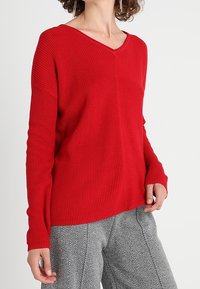 Pull en maille côtelée rouge avec un col en V et des manches longues, associé à un pantalon à motif pied-de-poule gris.