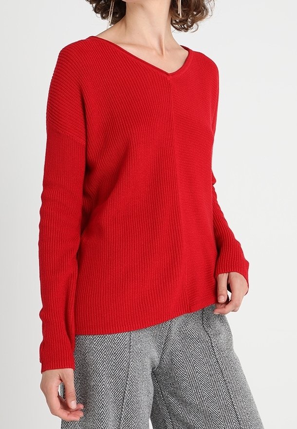 Pull en maille côtelée rouge avec un col en V et des manches longues, associé à un pantalon à motif pied-de-poule gris.