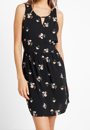 Černé šaty bez rukávů s růžovým a bílým květinovým vzorem, výstřihem ve tvaru klíčové dírky a řaseným pasem, které nosí žena.