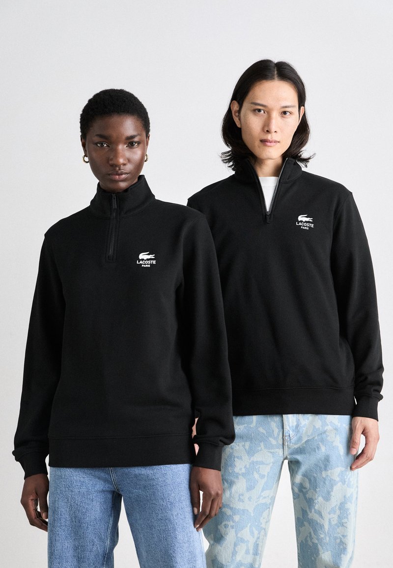 Juodi zipper tipo džemperiai su aukštu apykakle, puošiami baltais išsiuvinėti Lacoste logotipais. Dėvimi su šviesiai mėlynomis raštuotomis džinsinėmis kelnėmis.