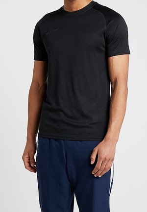 Homme portant un t-shirt de sport noir ajusté à manches courtes et un pantalon de sport bleu marine avec des bandes blanches sur les côtés.