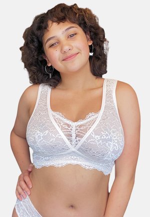Junge Frau mit lockigem Haar, die einen weißen Spitzen-Bralette und passende Unterwäsche trägt, lächelnd mit der Hand auf der Hüfte vor einfachem Hintergrund.