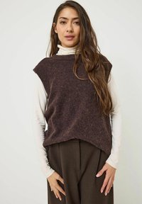 Pull en maille sans manches marron avec un col rond et un tissu texturé. Porté par-dessus un haut à manches longues blanc et associé à un pantalon marron.
