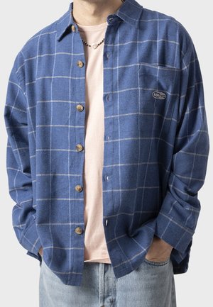 Man met een blauw geruit overhemd over een lichtroze t-shirt, lichtblauwe spijkerbroek, handen in de zakken en een kralenketting.