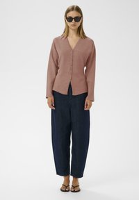 Rosa Button-Down-Cardigan mit V-Ausschnitt und langen Ärmeln, kombiniert mit lockeren dunklen Jeanshosen und Flip-Flops. Leichte Struktur sichtbar.