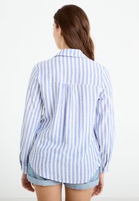 Mujer con cabello largo y castaño, vistiendo una camisa de manga larga con rayas verticales azul claro y blanco, y shorts de mezclilla, de espaldas.