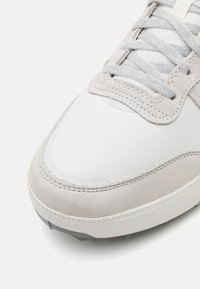 Sapatilha atlética de couro branco e camurça cinza com bico arredondado liso, atacadores texturizados e detalhes cinzas na lingueta e no colarinho.