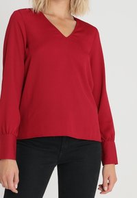 Rød V-halsbluse med lange ærmer og knaplukninger ved håndleddene, der har en glat tekstur og en afslappet pasform, parret med sorte bukser.