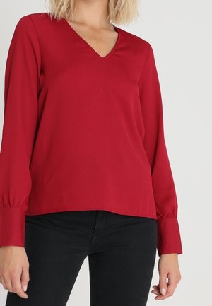 Röd V-ringad blus med långa ärmar och knäppta manschetter, som har en slät textur och avslappnad passform, kombinerad med svarta byxor.