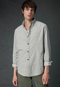 Cămașă gri deschis, cu un model subtil, nasturi negri și un mic logo pe pieptul stâng; asortată cu pantaloni verzi măslinii.