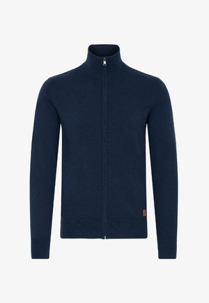 Mornarsko modra zip-up jopica, narejena iz mehke pletene tkanine, z visokim ovratnikom in rebrastimi manšetami. Vključuje majhno oznako z logotipom na robu.