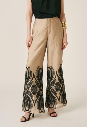 Mujer con pantalones beige de pierna ancha con bordado negro ornamentado en el bajo, top negro, brazalete dorado y sandalias negras de tacón.