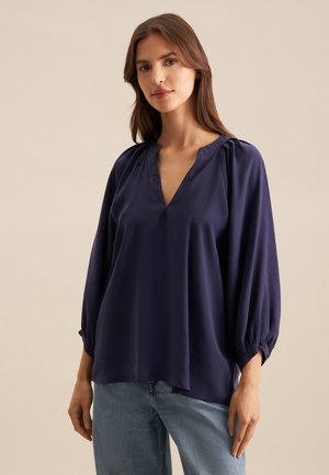 Blusa blu navy con scollo a V, design morbido e maniche a sbuffo, realizzata in tessuto liscio con una leggera lucentezza. Abbinata a jeans chiari.