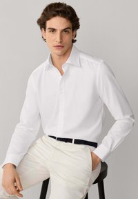 Jeune homme aux cheveux bruns ondulés portant une chemise blanche boutonnée et un pantalon clair, assis sur un tabouret devant un fond uni.