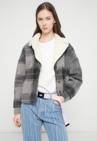 Jachetă gri cu carouri, cu guler din blană artificială crem, închidere cu nasturi și croială lejeră, asortată cu blugi drepți cu dungi albastre deschise și o curea roz.