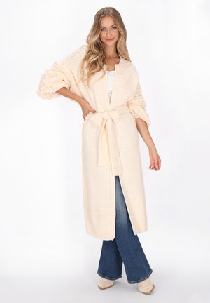 Long, knitted cardigan in cream with oversized sleeves and a self-tie belt. Worn over a white top and paired with wide-leg jeans.