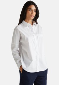 Camicia bianca a maniche lunghe con colletto bottonato, dettagli con taschino frontale e vestibilità rilassata, realizzata in tessuto liscio. Indossata con pantaloni scuri.