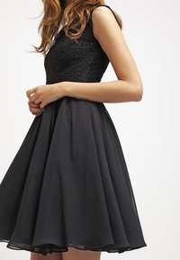 Robe noire avec un corsage ajusté en tissu texturé et une jupe en chiffon évasée de longueur genoux. Modèle sans manches, ourlet lisse.
