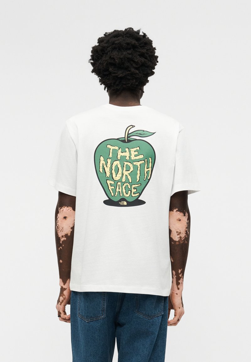 Άτομο με σκούρα σγουρά μαλλιά και λεύκη που φοράει ένα λευκό T-shirt με γραφικό πράσινο μήλο και το κείμενο «THE NORTH FACE» στην πλάτη.