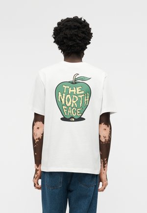 Άτομο με σκούρα σγουρά μαλλιά και λεύκη που φοράει ένα λευκό T-shirt με γραφικό πράσινο μήλο και το κείμενο «THE NORTH FACE» στην πλάτη.