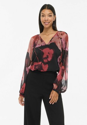 Une femme souriante porte un chemisier noir transparent à imprimé floral rose et à manches longues, assorti à un pantalon noir, sur fond blanc.