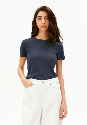 Femme aux longs cheveux foncés portant un t-shirt côtelé bleu marine à manches courtes et un pantalon blanc taille haute, une main dans la poche.