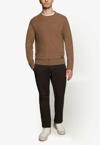Brun strikket sweater med rund hals, ribbede ærmer og bund. Kombineret med mørke bukser og lyse sneakers. Minimal brandingdetalje synlig.