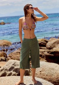 Bikini-top med lilla og mønstrede design, parret med løse, grønne culotter. Står på klipper ved stranden, barfodet, og ser ud over havet.